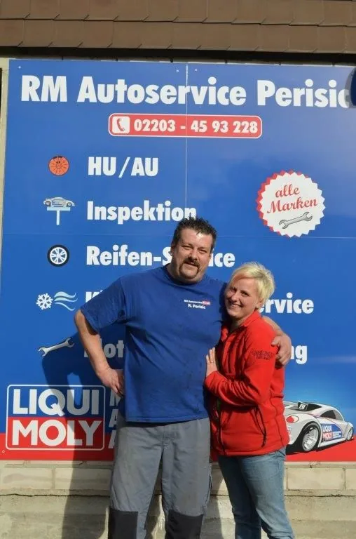 Das Inhaberpaar von RM Autoservice Perisic vor dem Werkstattschild mit Telefonnummer und Leistungsübersicht