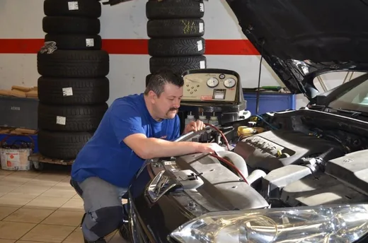 Klimaservicegerät steht am geöffneten Motorraum, ein Mitarbeiter schließt die Serviceschläuche an